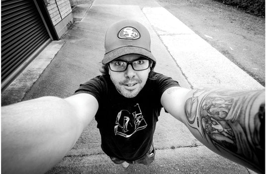 Person taking a selfie on a sidewalk wearing a cap and t-shirt.