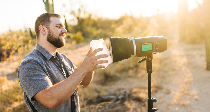 A bearded man in a gray shirt stands outdoors in a desert setting, holding a white MagMod diffuser in front of a large light on a stand. Warm golden sunlight shines from the background.